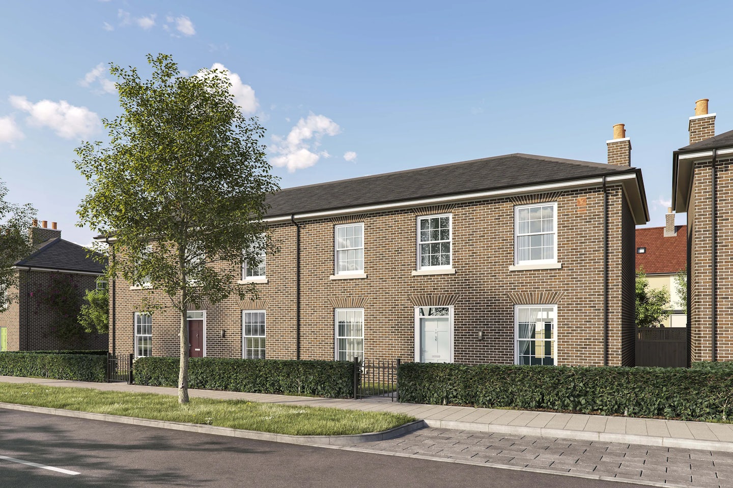 Row of modern brick houses with pitched roofs and white-framed sash windows along a paved street. Each house has a front door, some with small hedges and railings in front. A young tree is planted on the pavement in the foreground, and the sky is bright blue with scattered clouds.