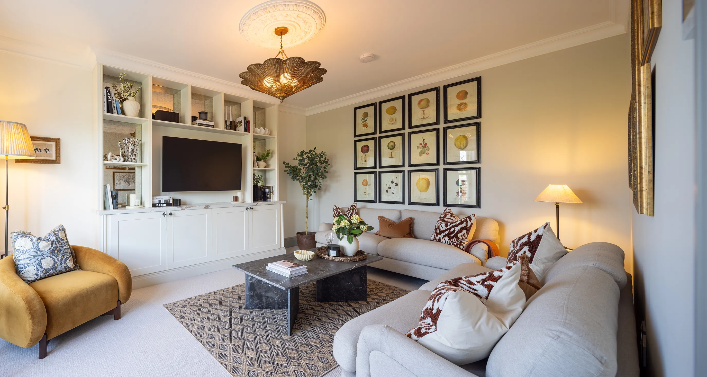 Living room with a modern design. A large built-in shelving unit frames a flat-screen TV, surrounded by books and decorative items. Two sofas—one beige, one light gray—are adorned with patterned cushions, complemented by a mustard-coloured armchair. A coffee table sits atop a patterned rug, holding books and decor. Framed botanical prints in a grid pattern decorate the walls. Various lighting sources, including a ceiling fixture, floor lamp, and table lamp, create a warm ambiance. A potted plant adds greenery to the inviting space.