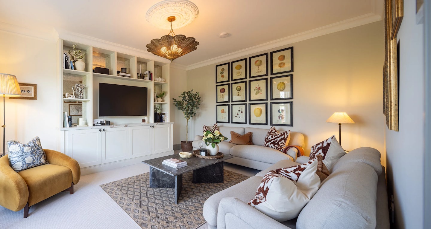 Living room with a modern design. A large built-in shelving unit frames a flat-screen TV, surrounded by books and decorative items. Two sofas—one beige, one light gray—are adorned with patterned cushions, complemented by a mustard-coloured armchair. A coffee table sits atop a patterned rug, holding books and decor. Framed botanical prints in a grid pattern decorate the walls. Various lighting sources, including a ceiling fixture, floor lamp, and table lamp, create a warm ambiance. A potted plant adds greenery to the inviting space.