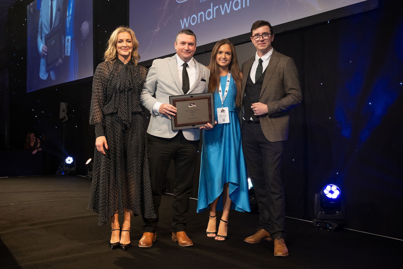 Four people standing on a stage at an awards event, dressed in formal attire, with one person holding a framed certificate. A large screen with event branding is visible in the background, and stage lights illuminate the scene.