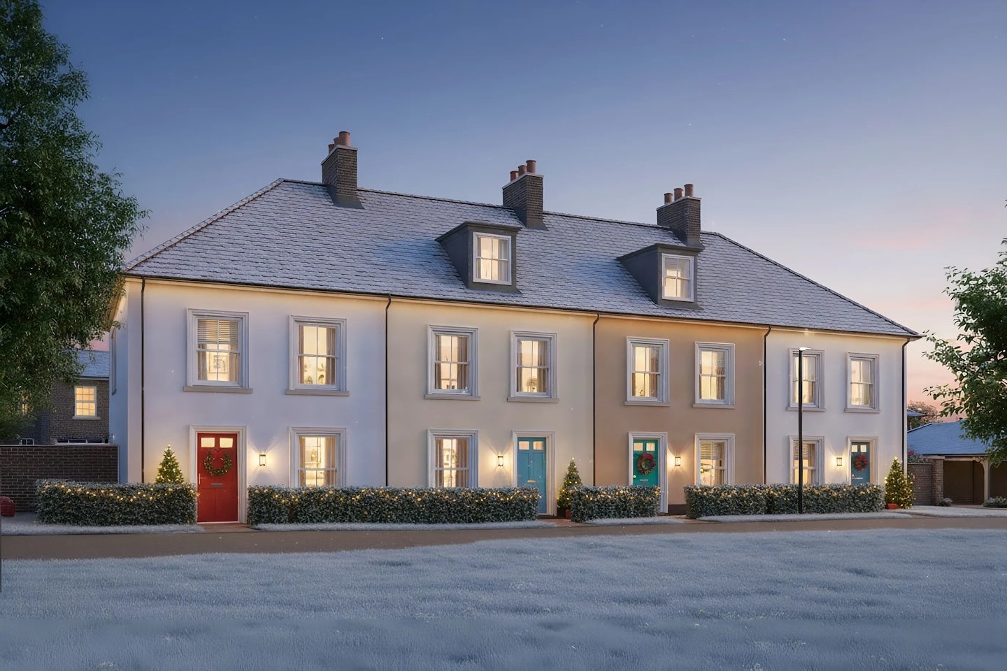 Row of elegant three-story townhouses with white façades and pitched roofs, decorated for Christmas with wreaths on the doors, illuminated shrubs, and small lit trees by the entrances. The photo is taken at dusk with warm interior lights glowing through the windows.