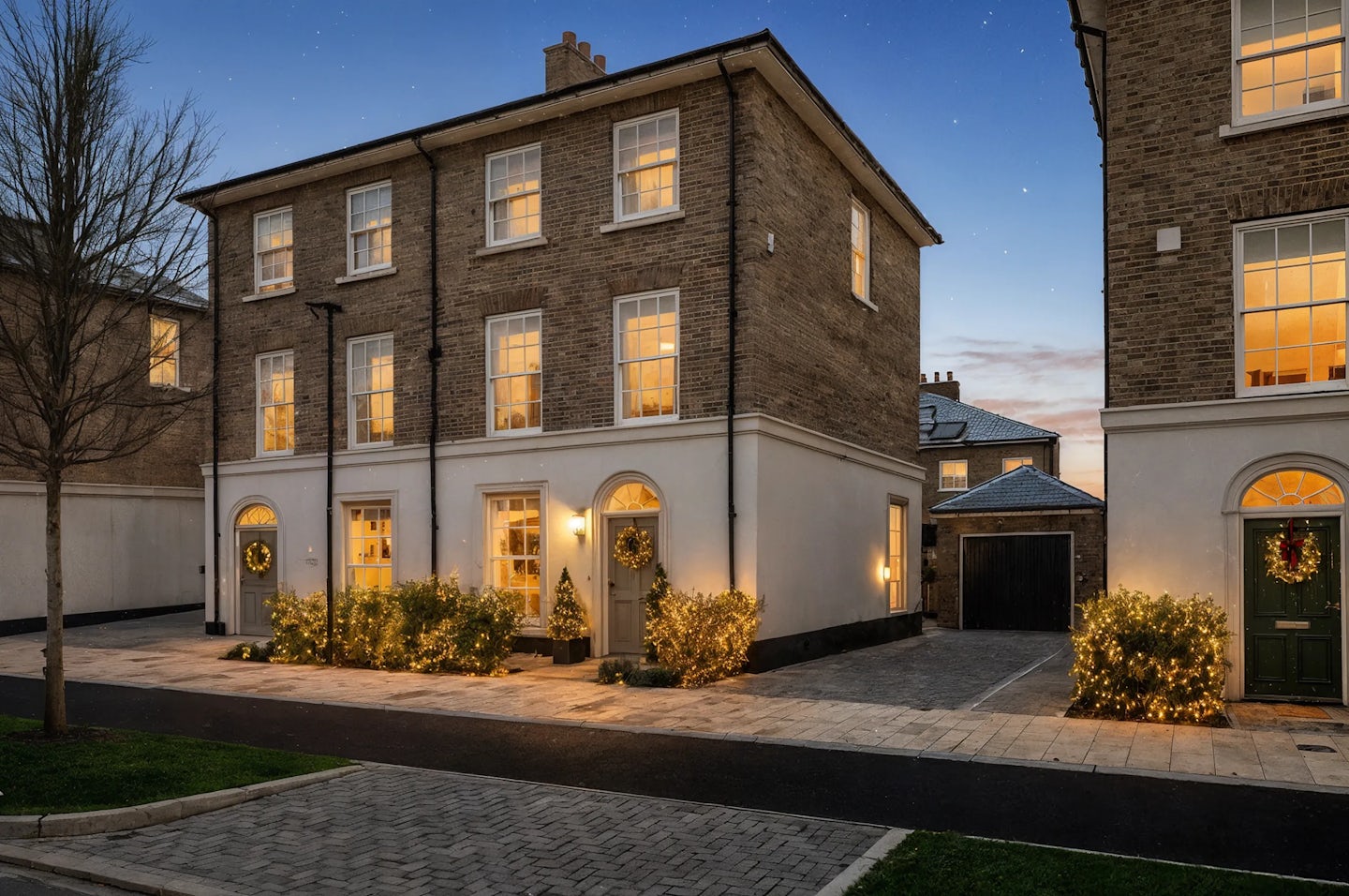 Elegant three-story brick townhouse decorated with warm white Christmas lights and wreaths on the doors, viewed at dusk. The property features large sash windows glowing from interior lighting, a paved driveway, and a small tree in the foreground.