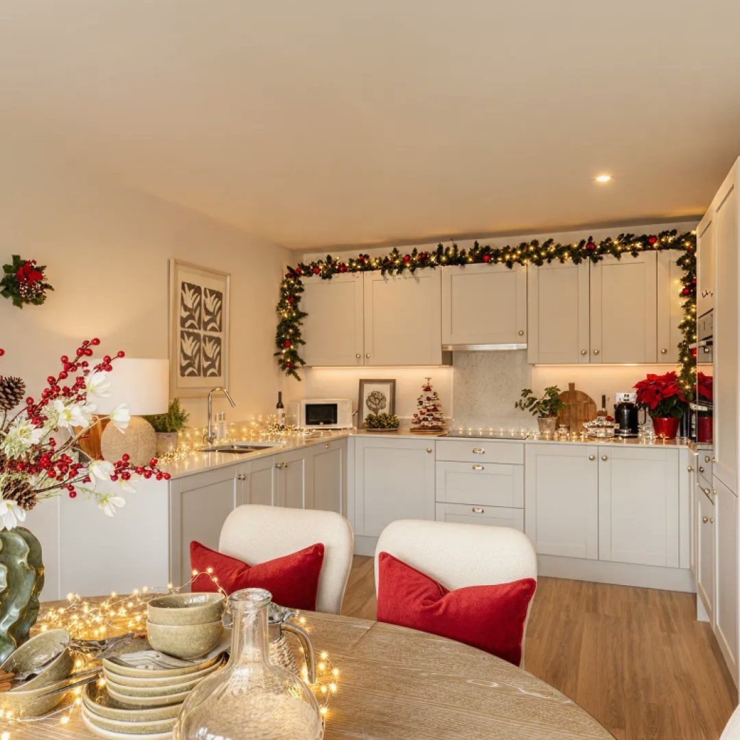Modern kitchen decorated for Christmas with white cabinets, wooden flooring, and a garland with red bows and lights draped above the units. A round dining table in the foreground is set with crockery, glassware, and festive accents, including red cushions and string lights.