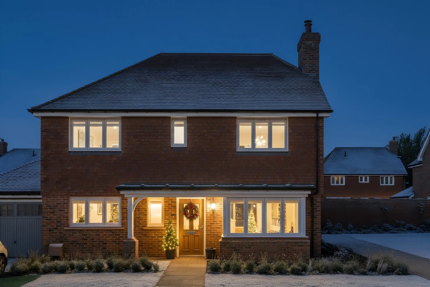 Two-story brick house decorated for Christmas with a wreath on the front door, a small illuminated tree by the entrance, and warm interior lights glowing through large windows. The photo is taken at dusk with a light frost on the driveway and roof.