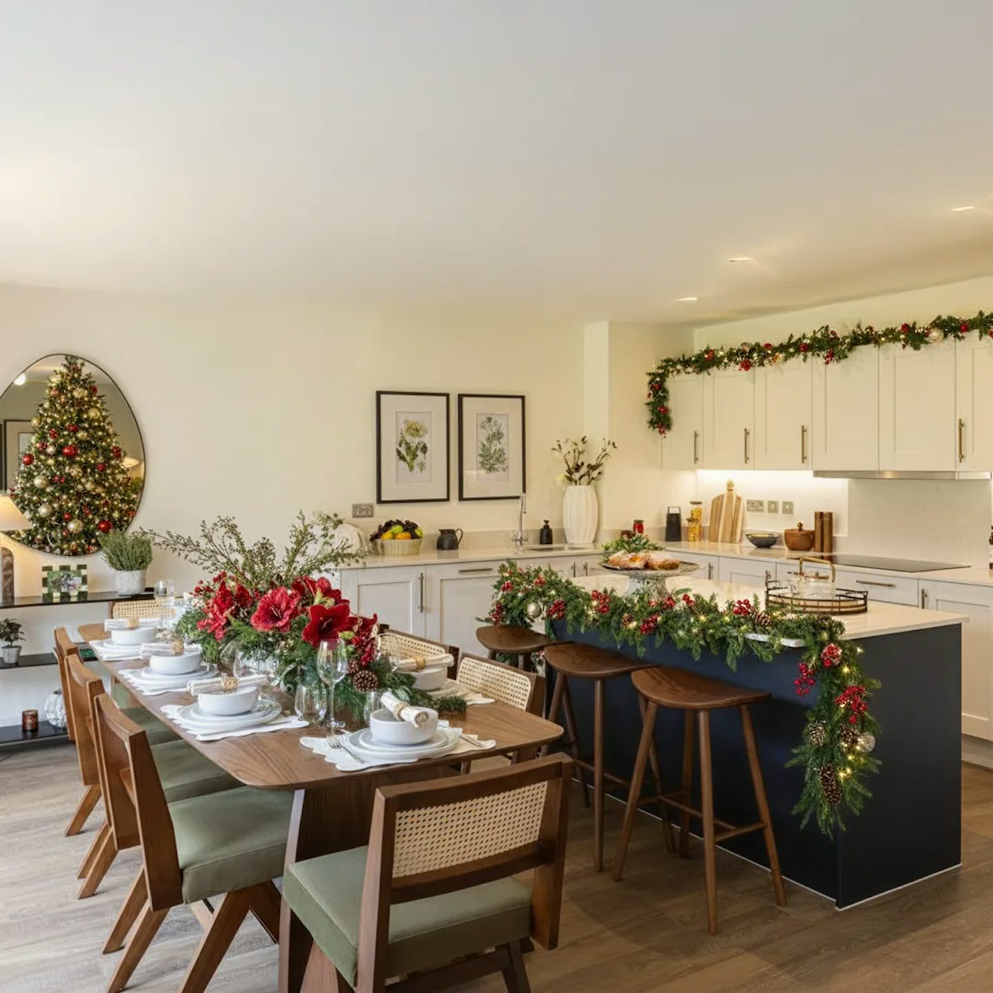 Open-plan kitchen and dining area decorated for Christmas with white cabinets, a dark island, and wooden flooring. The island and wall units are adorned with festive garlands, while the dining table is set with plates, glasses, and a centerpiece of red flowers and greenery. A decorated Christmas tree stands in the corner near a mirror.