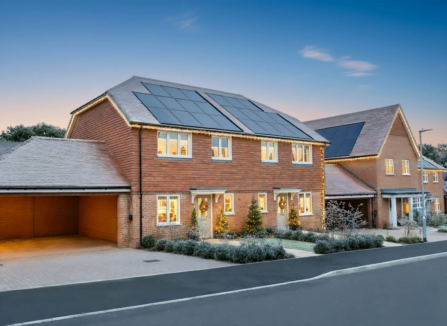 Modern two-story brick house with solar panels on the roof, decorated for Christmas with warm white lights along the roofline and around the porch. Wreaths hang on the doors, and small illuminated trees are placed at the entrance. The property has a paved driveway and landscaped shrubs, photographed at dusk.