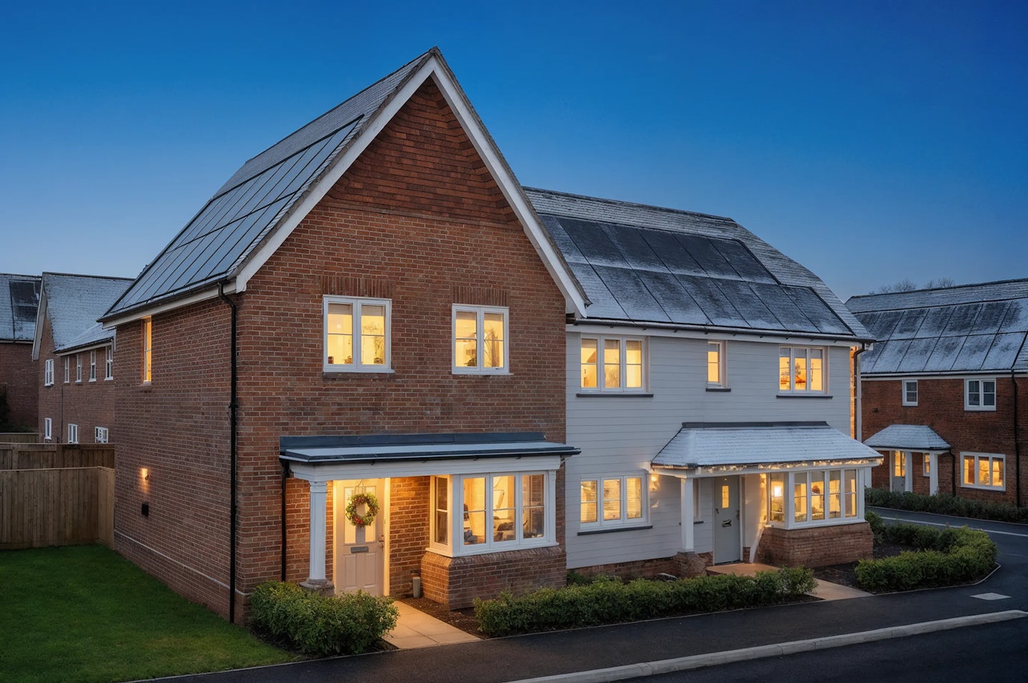 Two-story detached house with a mix of red brick and white cladding, decorated for Christmas with warm white lights along the roofline and a wreath on the front door. Solar panels are visible on the roof, and the property is photographed at dusk with glowing interior windows and a neat front garden.