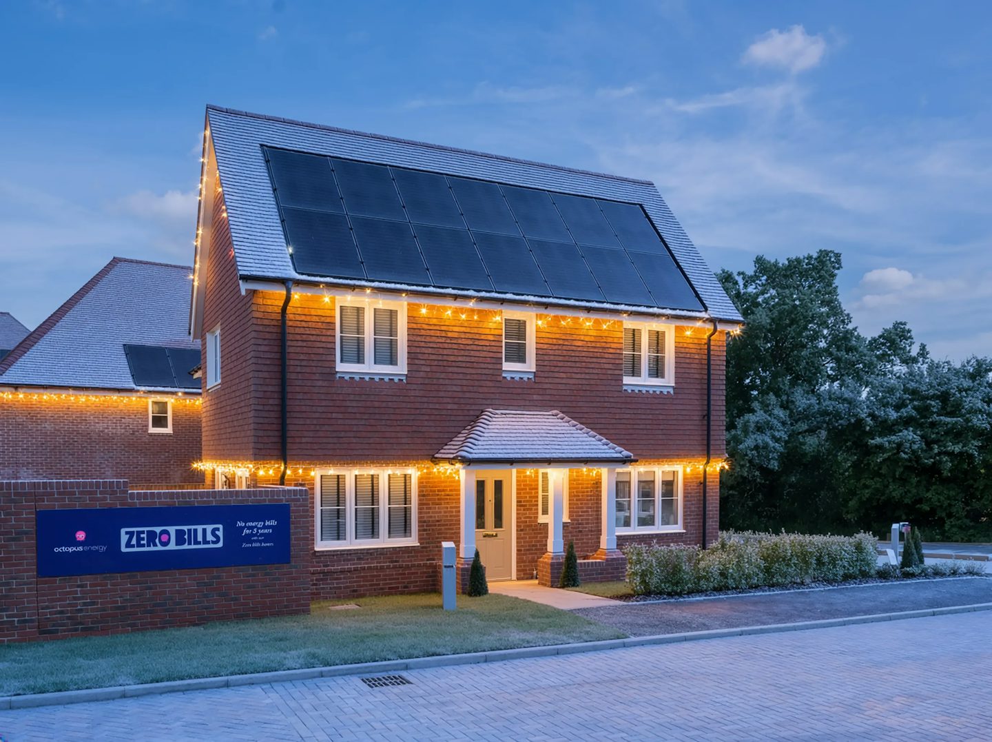 Modern two-story brick house with solar panels on the roof, decorated for Christmas with warm white lights along the roofline and around the porch. A sign in front reads “Zero Bills,” and the property is photographed at dusk with a paved driveway and landscaped shrubs.