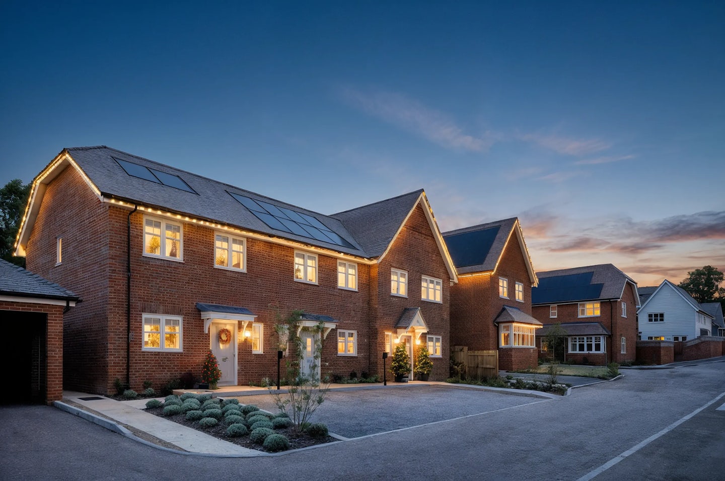 Row of modern brick houses with solar panels on the roofs, decorated for Christmas with warm white lights along the rooflines and wreaths on the doors. Small illuminated trees are placed at the entrances, and the photo is taken at dusk with a clear sky.