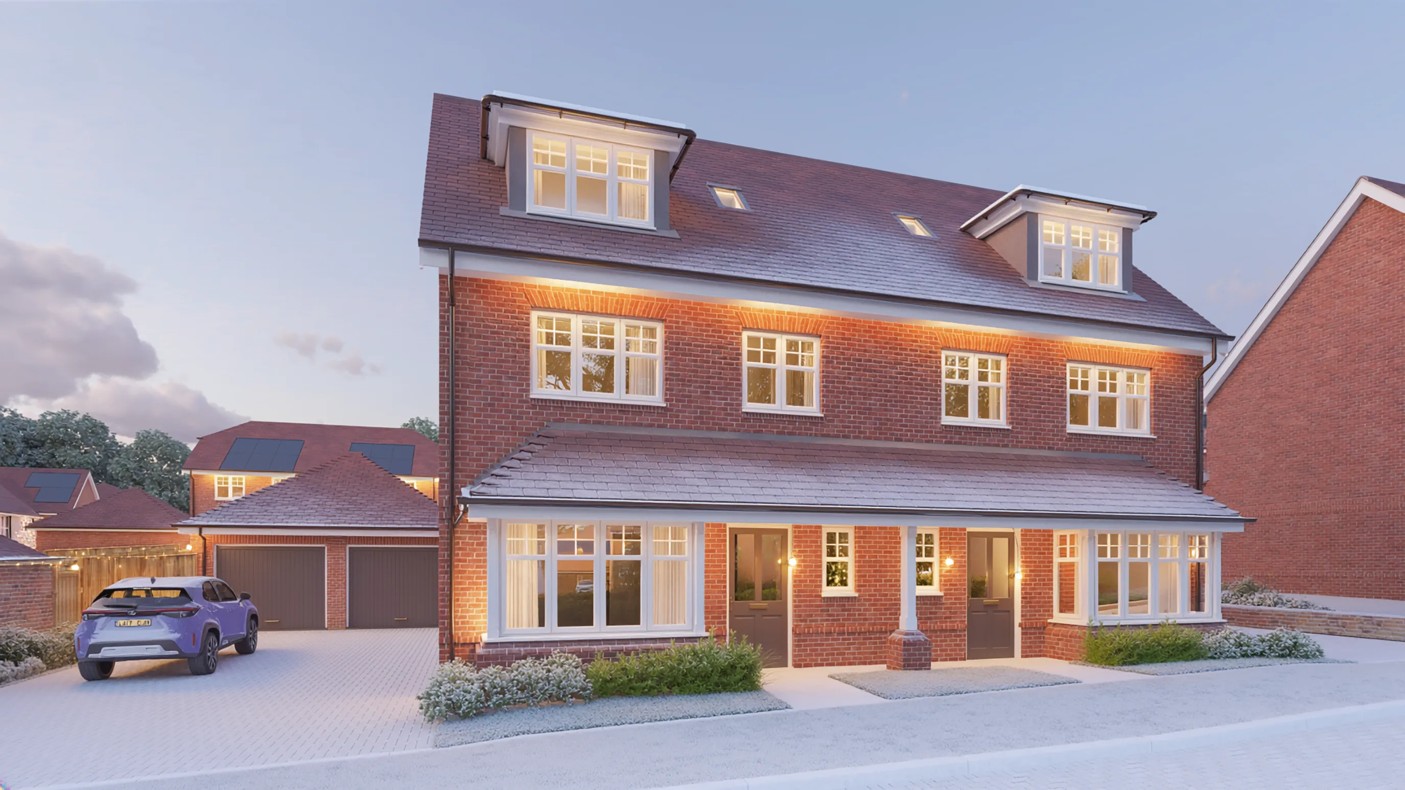 Three-story red-brick townhouse with dormer windows on the top floor, decorated for Christmas with warm white lights along the roofline and around the porch. The property has large sash windows, a paved driveway with a parked car, and landscaped shrubs in front, photographed at dusk.