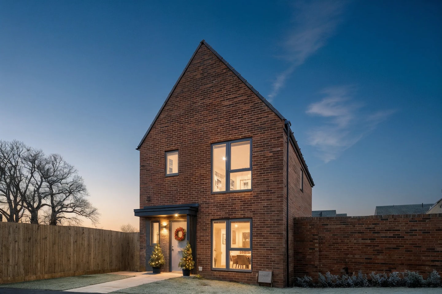 Two-story red-brick house with a steep pitched roof, decorated for Christmas with a wreath on the front door and two illuminated trees at the entrance. Warm interior lights glow through large windows, and the property is photographed at dusk with a clear sky and a frosty lawn.