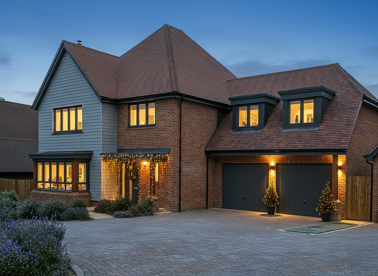 Large detached house with a mix of red brick and gray cladding, decorated for Christmas with warm white lights around the porch and two illuminated trees by the garage doors. The property features a pitched roof, dormer windows, and a wide paved driveway, photographed at dusk with glowing interior lights.