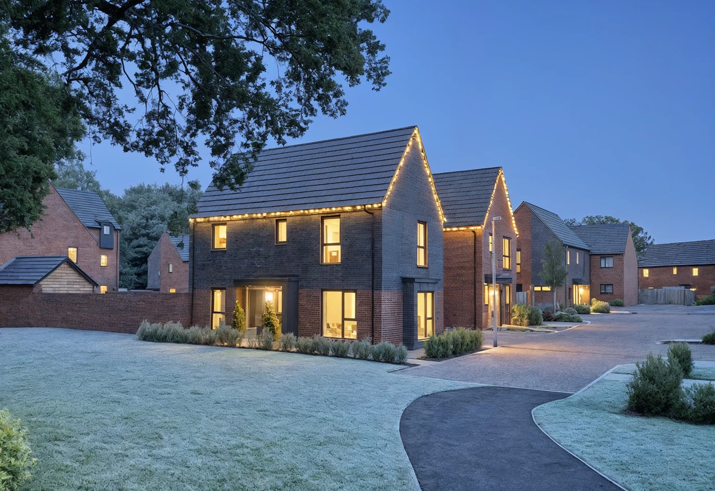 Row of modern two-story brick houses with pitched roofs, decorated for Christmas with warm white lights along the rooflines. The homes have large windows glowing from interior lights, landscaped front gardens, and a frosty driveway, photographed at dusk under a clear blue sky.