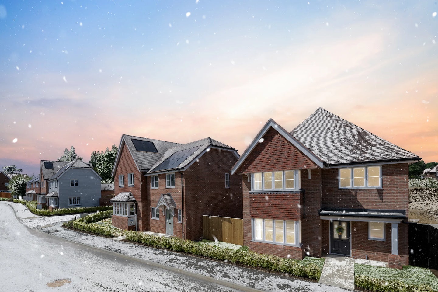 Row of modern two-story brick houses with pitched roofs, photographed at sunset with a frosty driveway and falling snow. The homes feature large windows glowing with warm interior light, landscaped front gardens, and one house has a wreath on the front door.