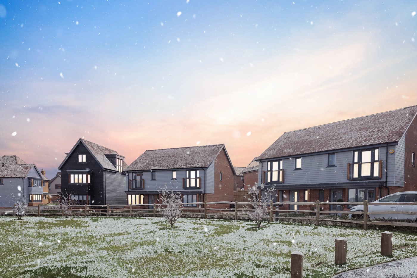 Row of modern two-story houses with a mix of red brick and gray cladding, viewed across a frosty green space with wooden fencing and small trees. Snowflakes are falling, and the photo is taken at sunset with a colorful sky.