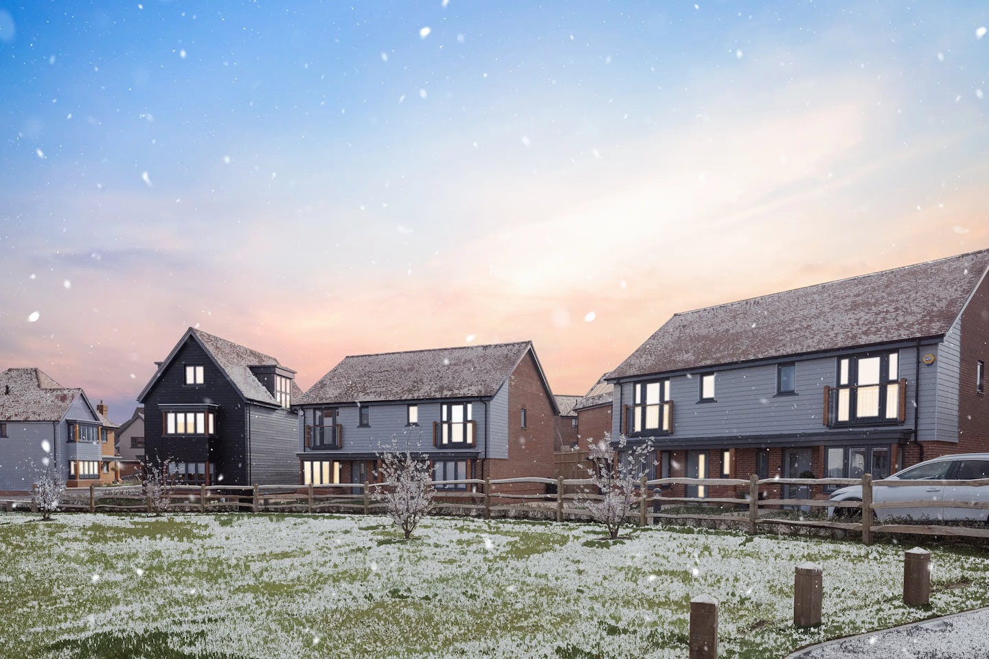 Row of modern two-story houses with a mix of red brick and gray cladding, viewed across a frosty green space with wooden fencing and small trees. Snowflakes are falling, and the photo is taken at sunset with a colorful sky.