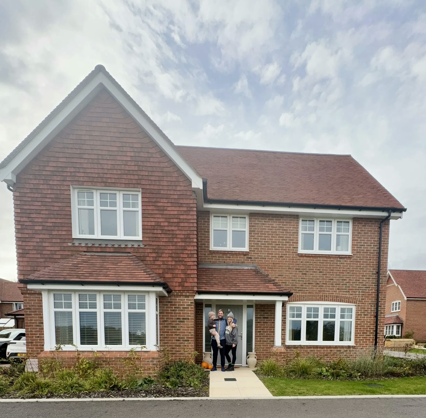 A two-story detached brick house with a red tiled roof and white-framed windows. Three people are standing together at the front door, with a small paved path leading to the entrance. Low greenery borders the house, and similar homes are visible in the background under a partly cloudy sky.