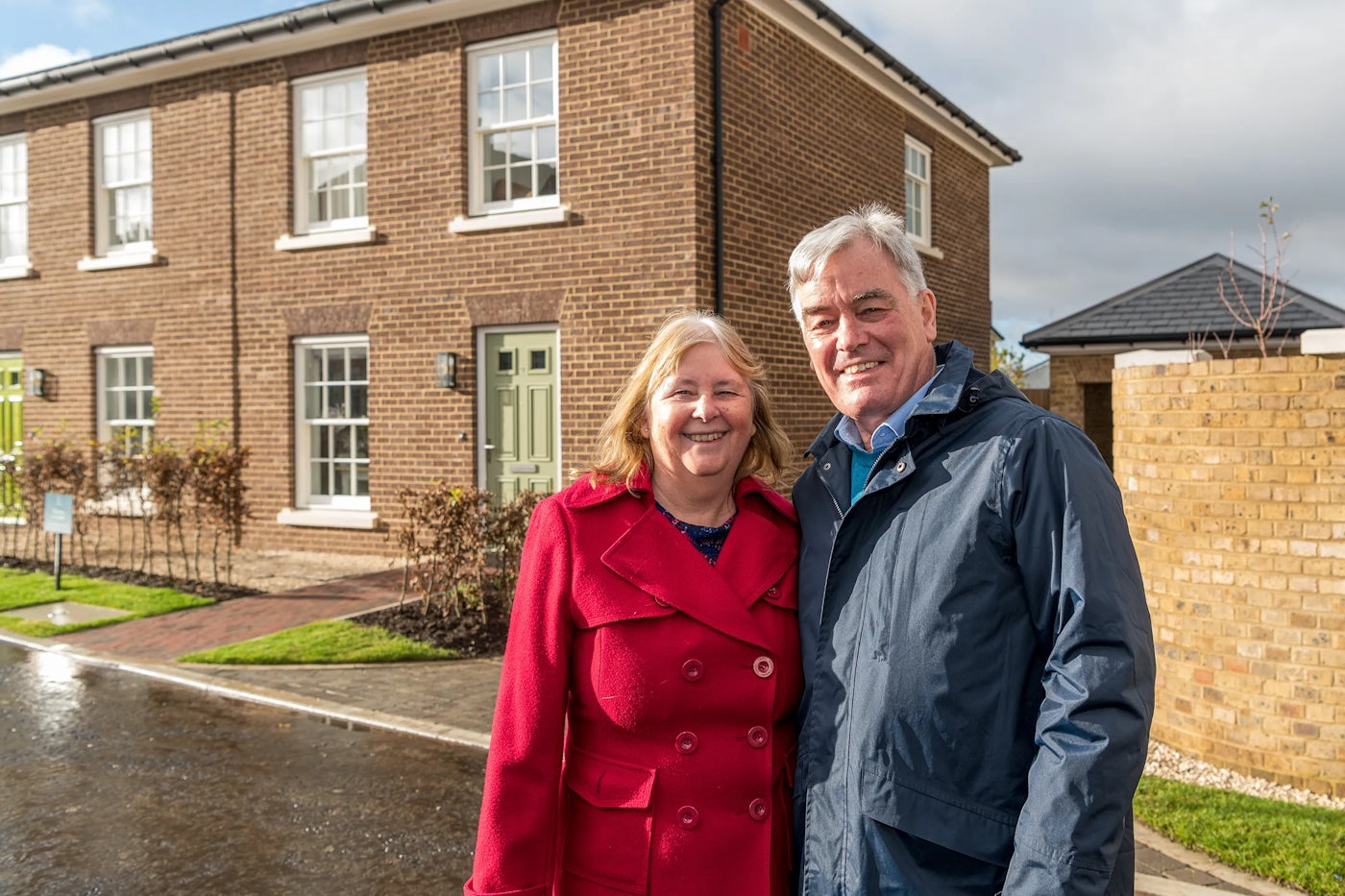 A couple standing together on a residential street in front of a newly built brick house on a bright day.