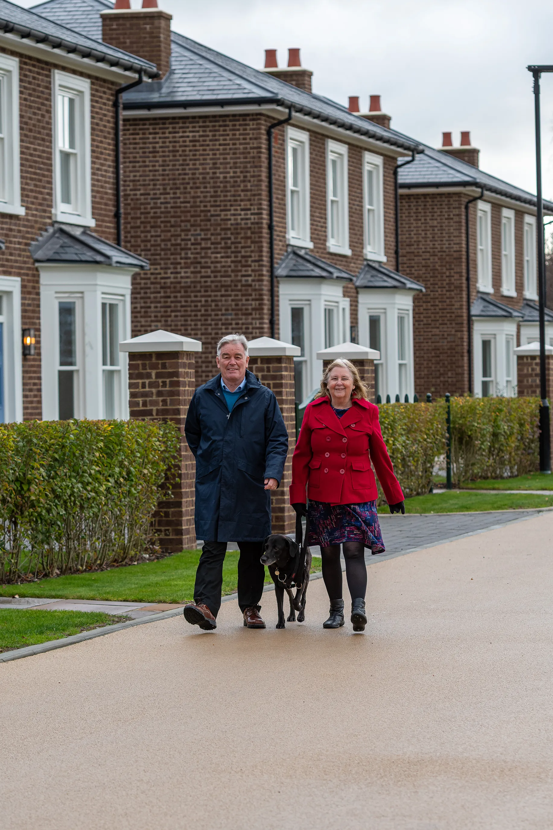 A couple walking a black dog along a tidy residential street with rows of new brick homes.