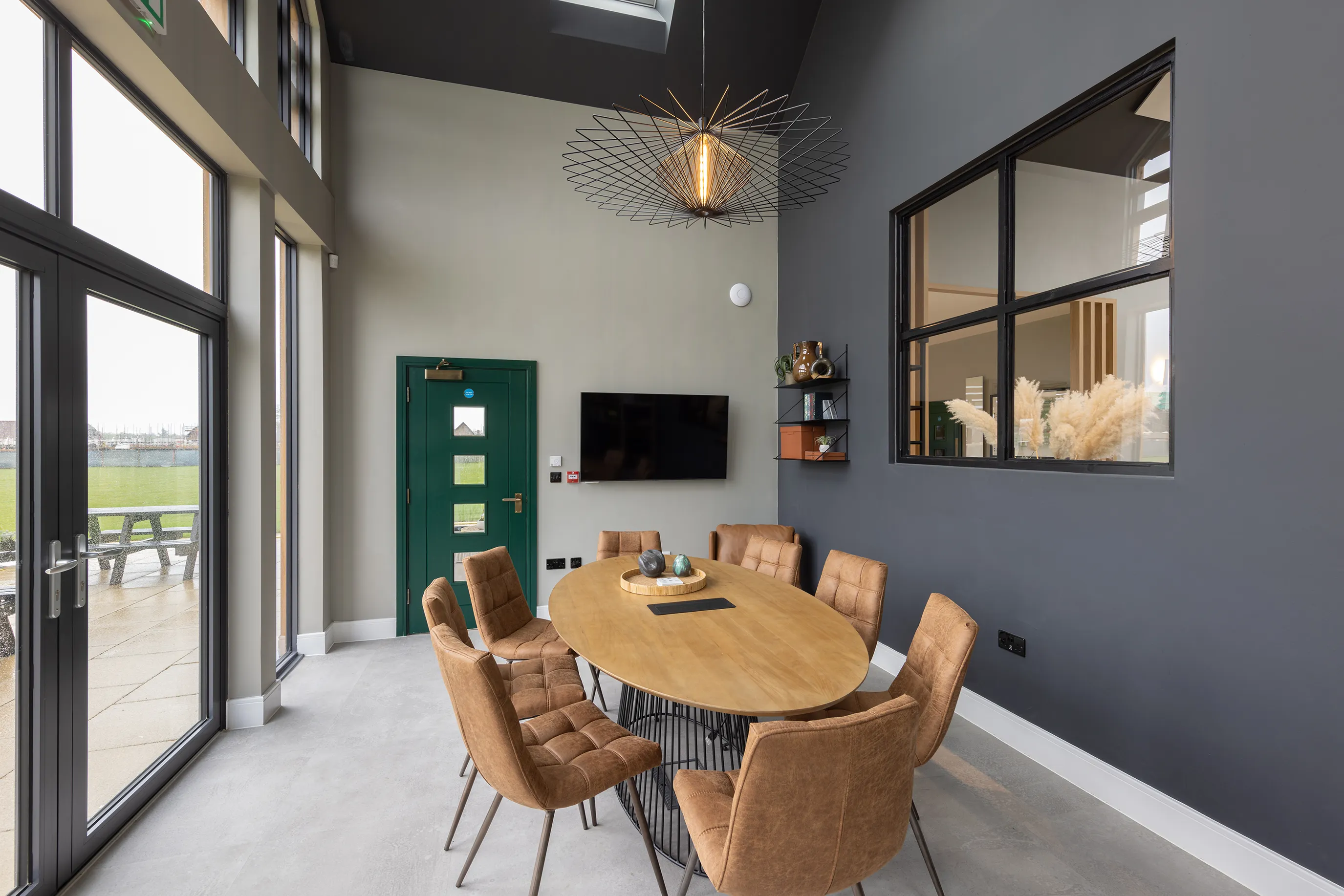 A modern meeting room with an oval wooden table, brown upholstered chairs, large windows, and a glass door leading outside.