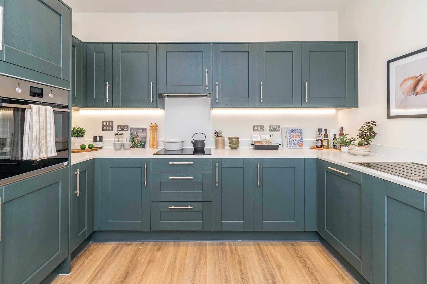 A modern kitchen with dark green cabinets, built‑in appliances, light worktops, and wooden flooring.