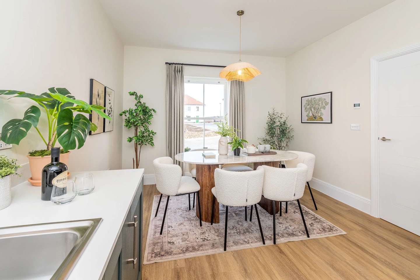 A dining area with a round table, six chairs, plants, wooden flooring, and a window letting in natural light.