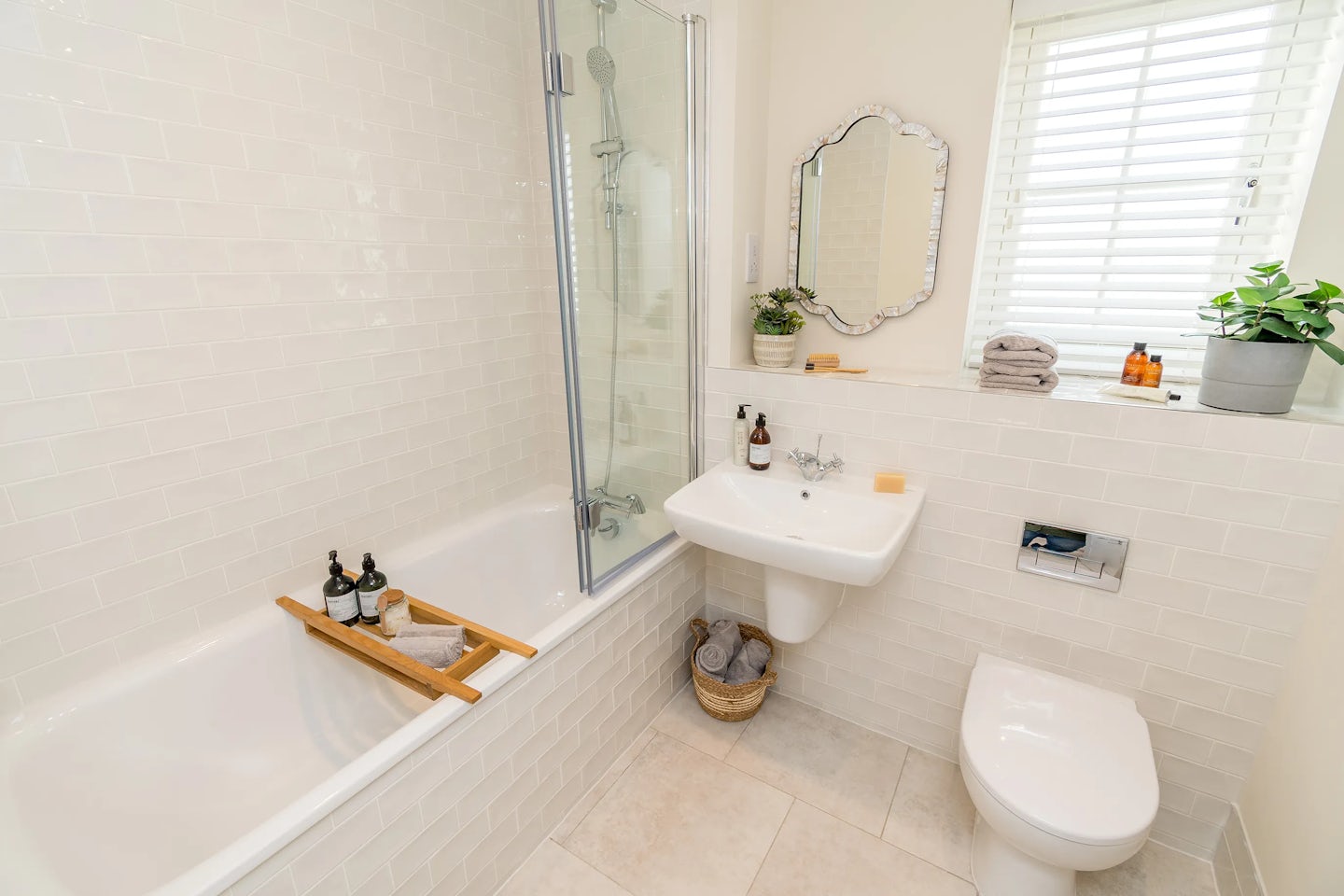 A bright bathroom with a bathtub and shower, white tiled walls, a wall‑mounted sink, a toilet, and a window letting in natural light.