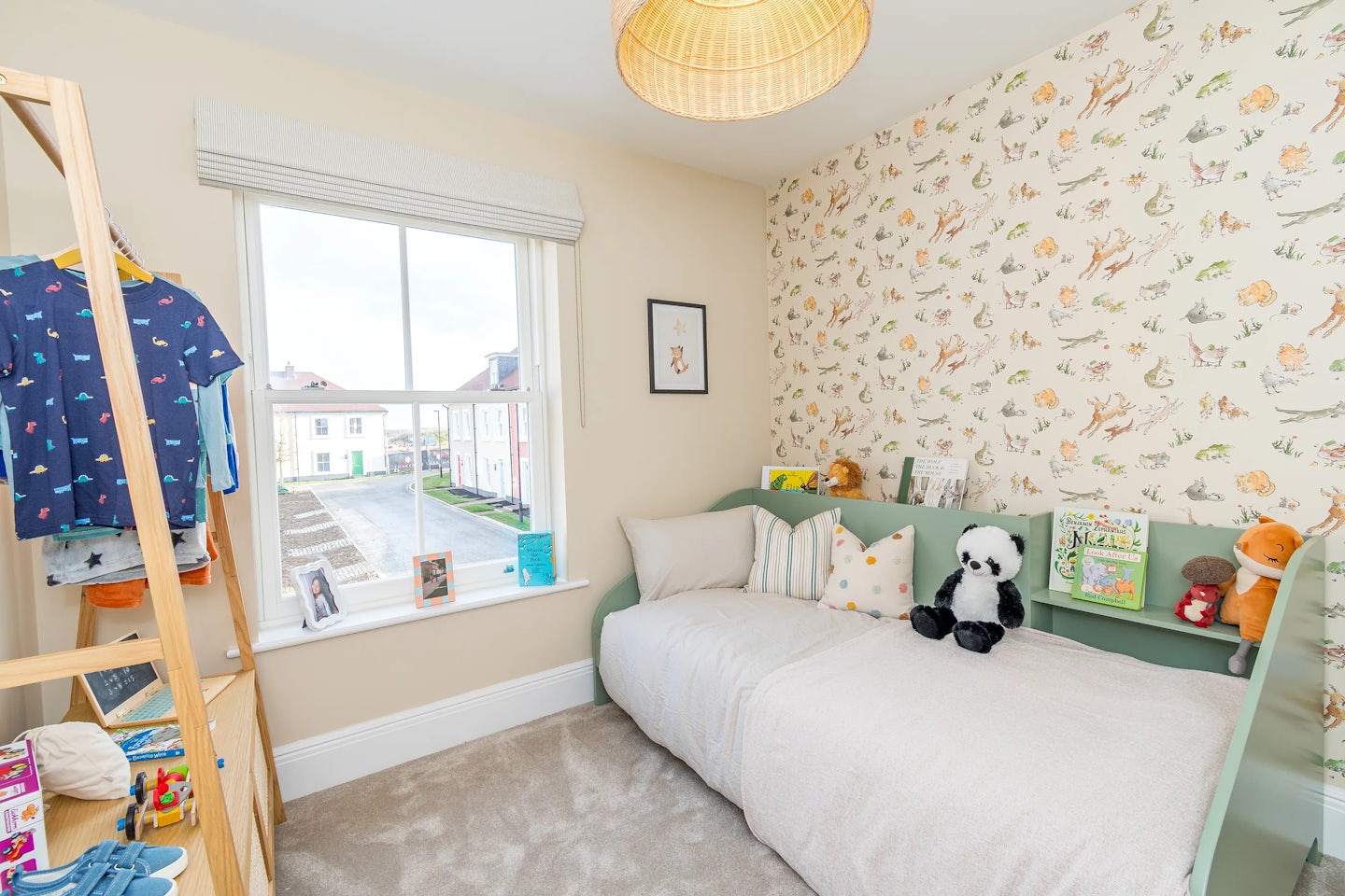 A child’s bedroom with a single bed, patterned wallpaper, soft toys on the bed, and a window overlooking nearby houses.