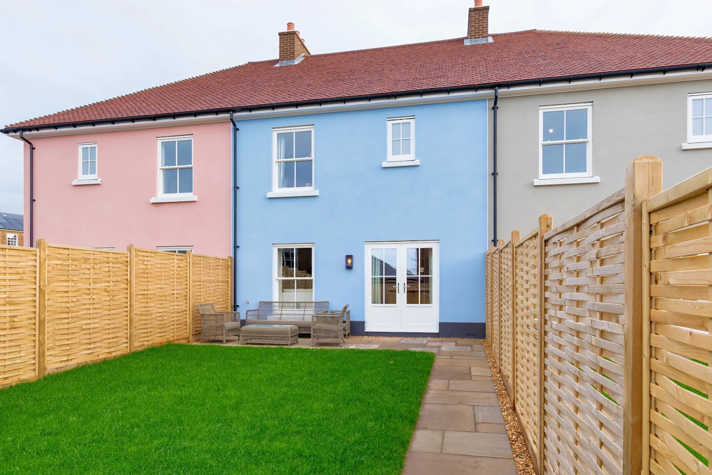 A fenced rear garden with a green lawn, paved patio, outdoor seating, and the back of a terraced house.