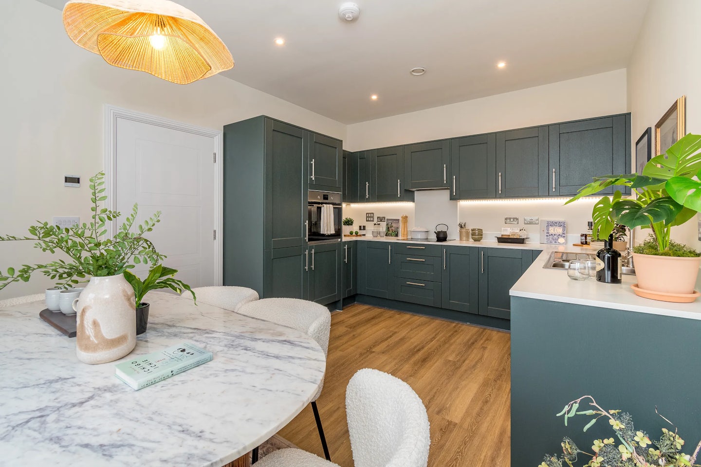A modern kitchen with dark green cabinets, a marble dining table, wooden flooring, and plants on the worktops.