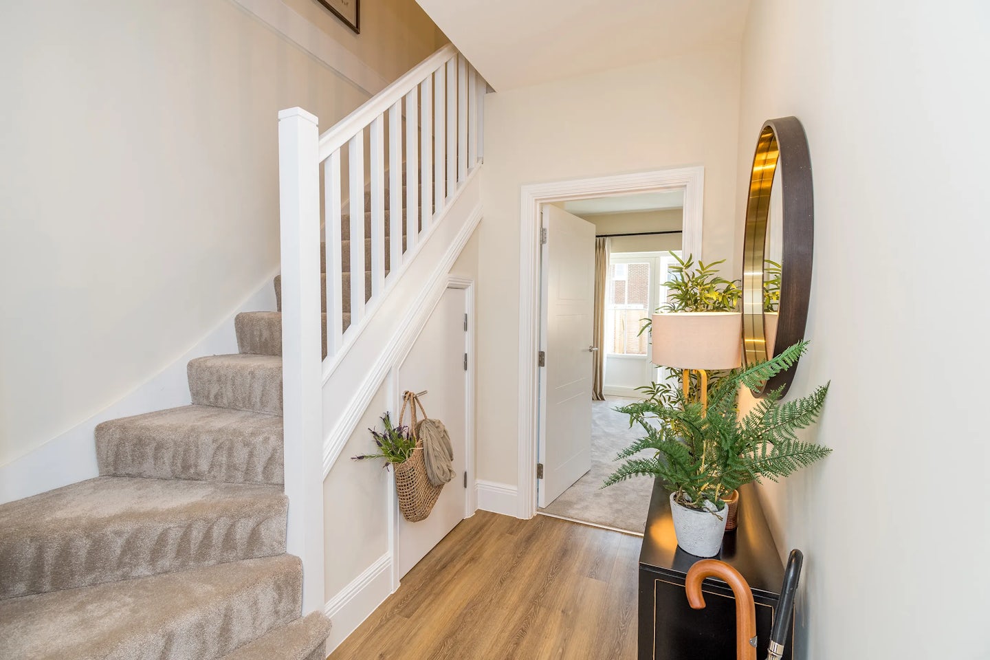A bright hallway with carpeted stairs, wooden flooring, a console table with plants, and a doorway leading to another room.
