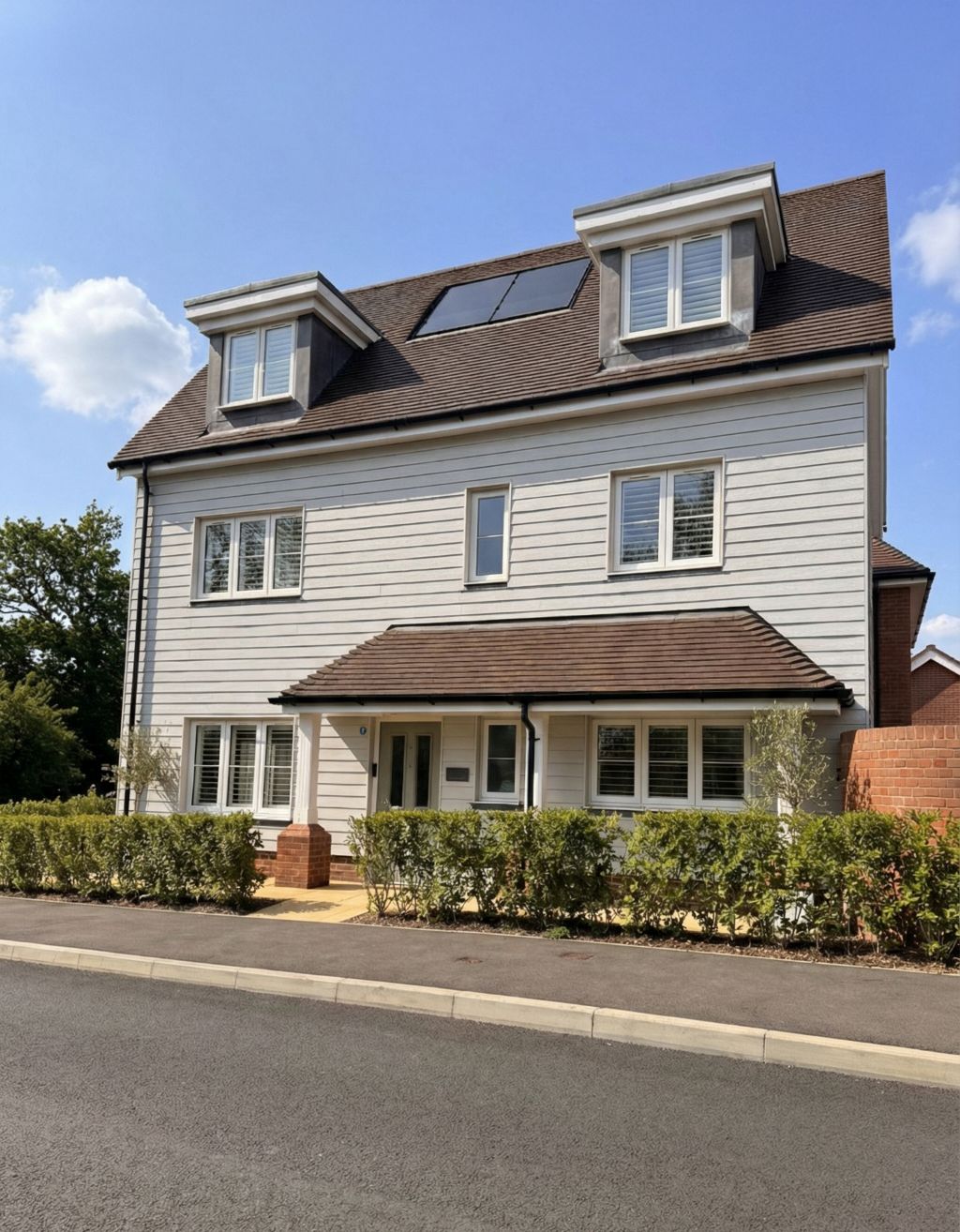 Front view of a modern three‑storey detached house with light grey cladding, dormer windows, and a small hedge along the pavement