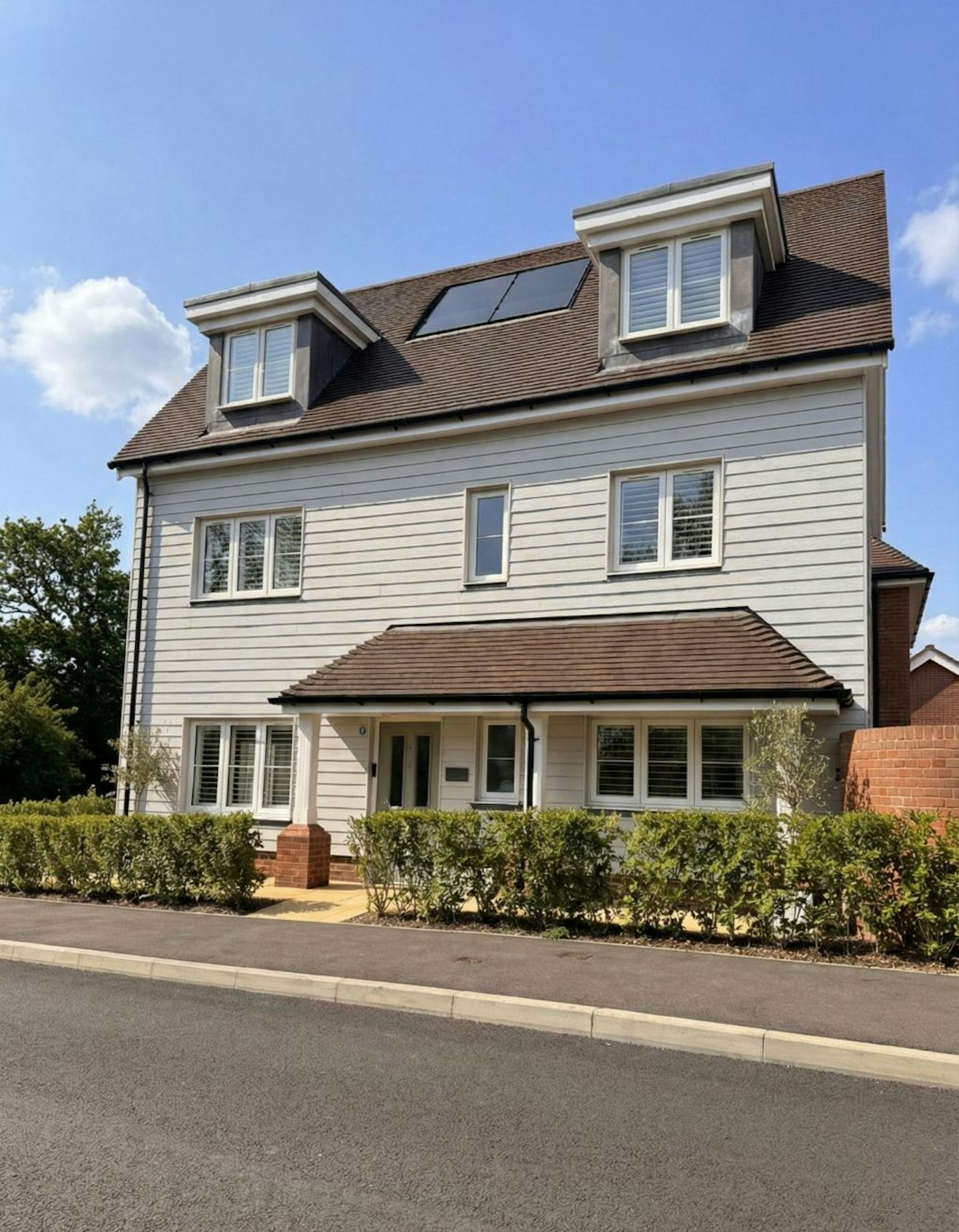 Front view of a modern three‑storey detached house with light grey cladding, dormer windows, and a small hedge along the pavement