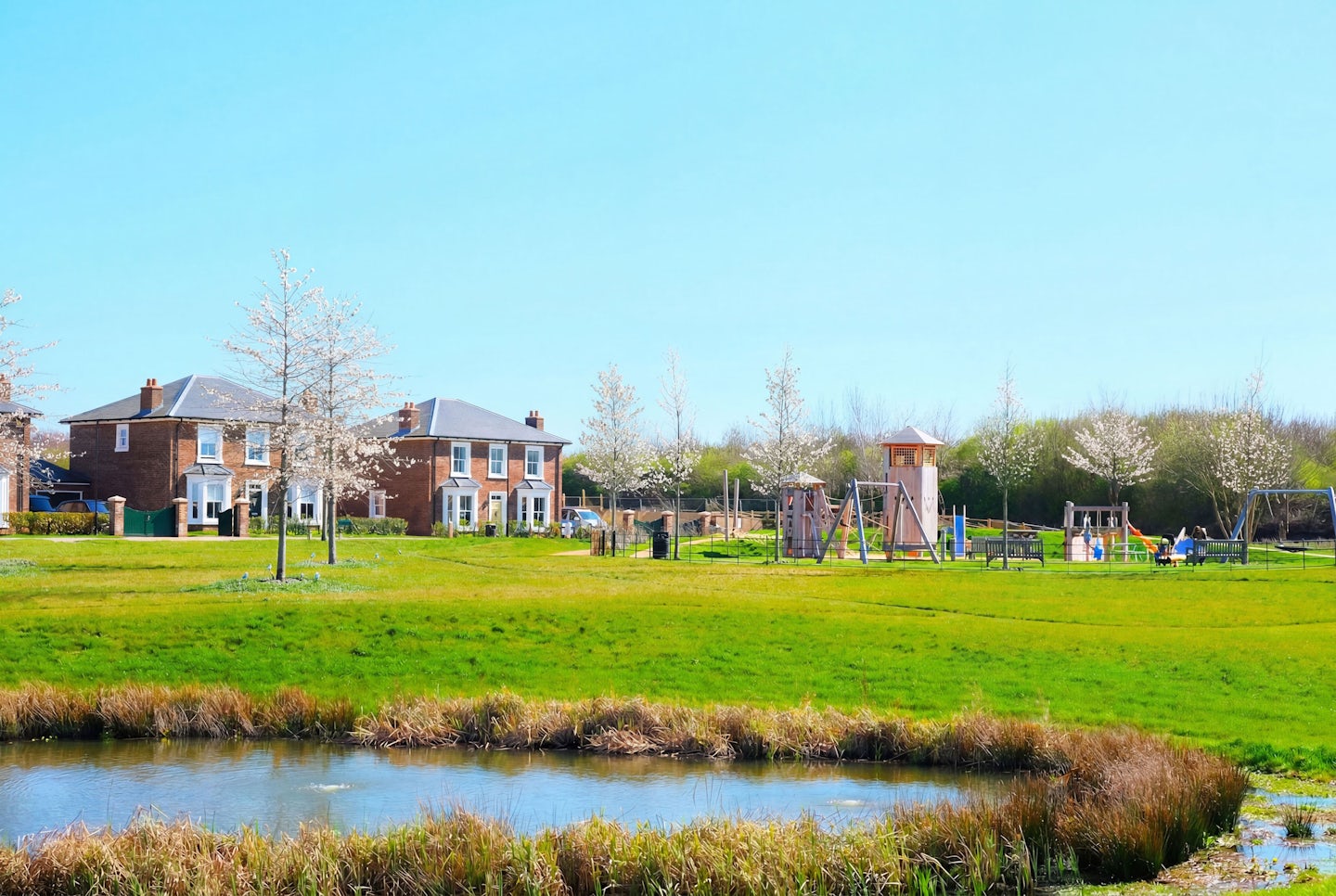 Green open space with a small pond, blossom trees, a children’s play area, and new homes in the background.