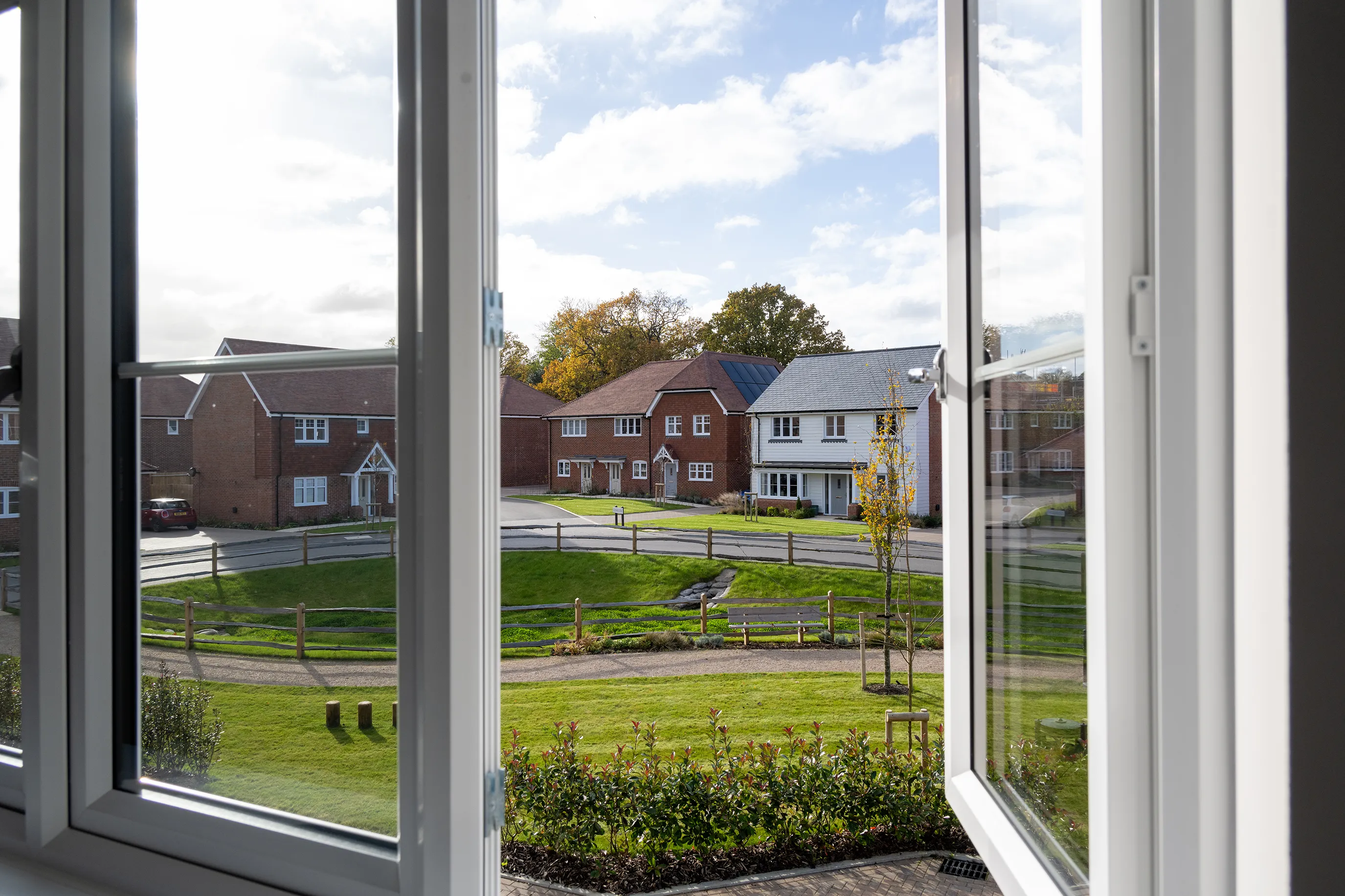 View through an open window looking onto a residential street with houses, grass, and trees.