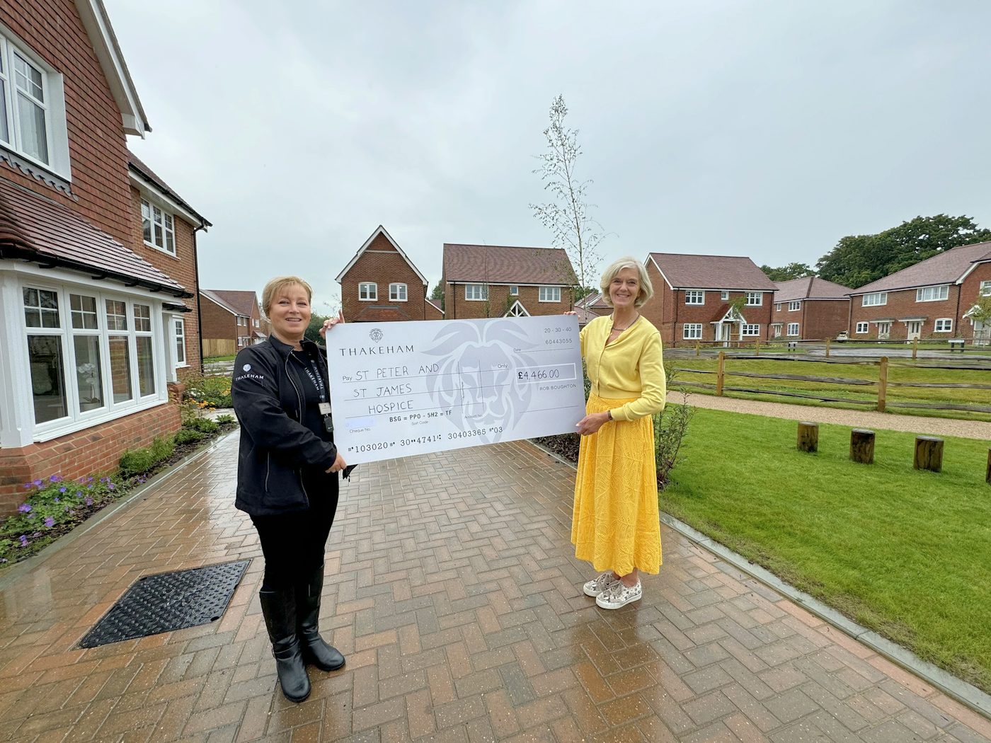 Two people standing on a paved path in a new housing development, holding a large charity cheque, with red‑brick homes and green open space behind them.