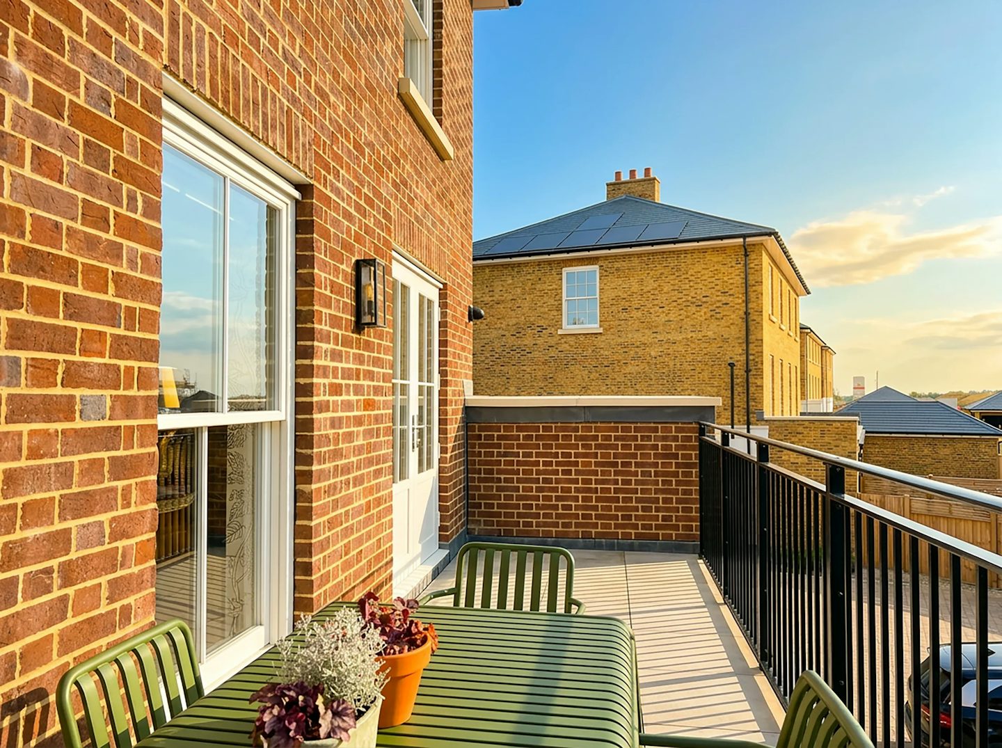 Balcony terrace with outdoor seating, brick walls and black metal railing, with nearby houses in the background.
