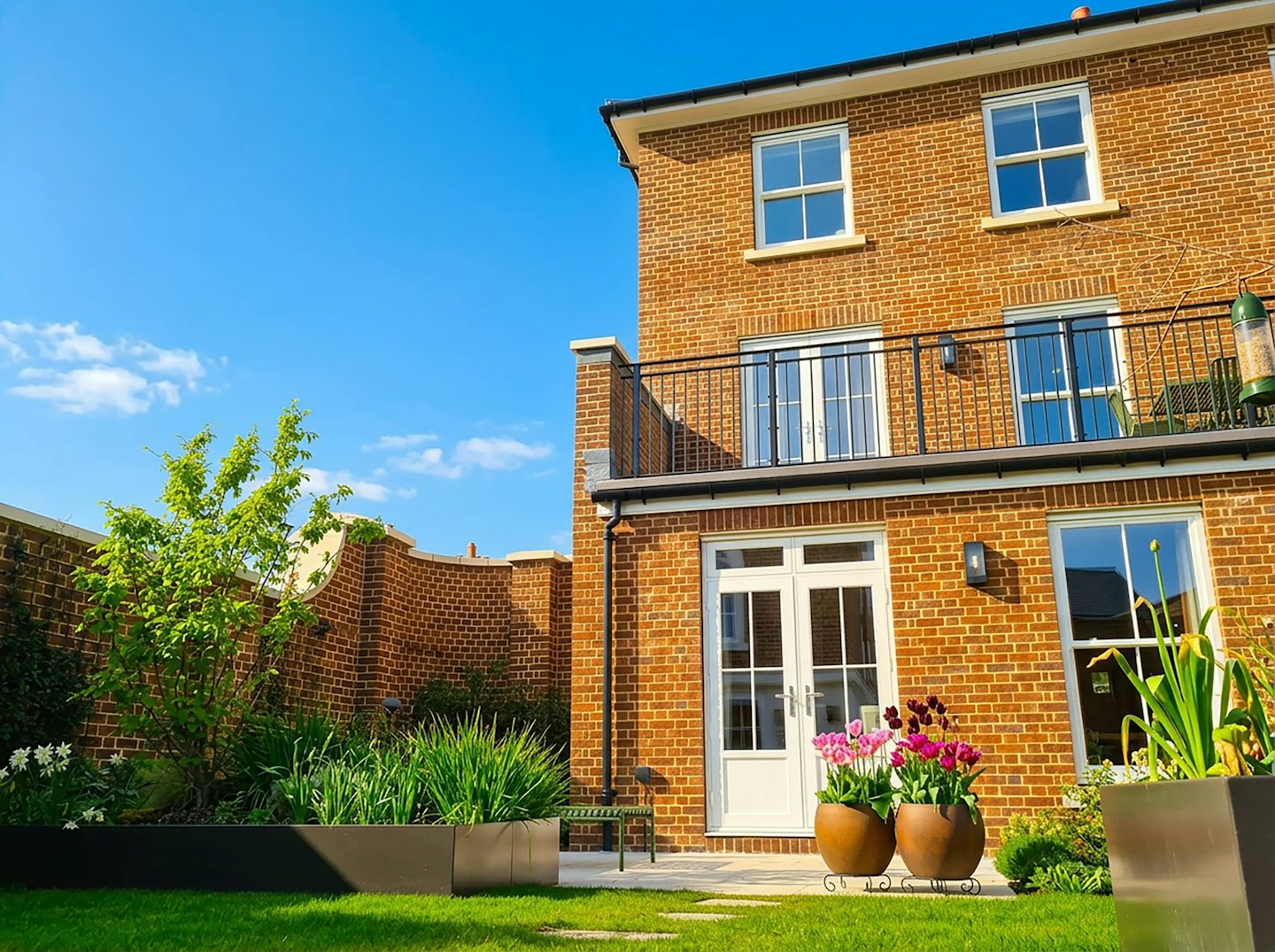 Rear view of a brick house with patio doors, first-floor balcony, paved garden, and planted borders.