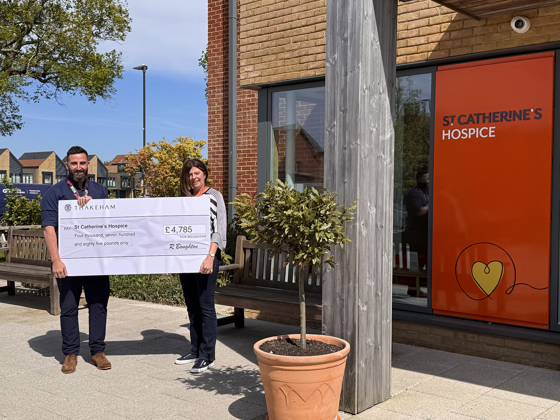 Two people holding a donation cheque for St Catherine’s Hospice outside a hospice building, with an orange hospice sign on the wall.