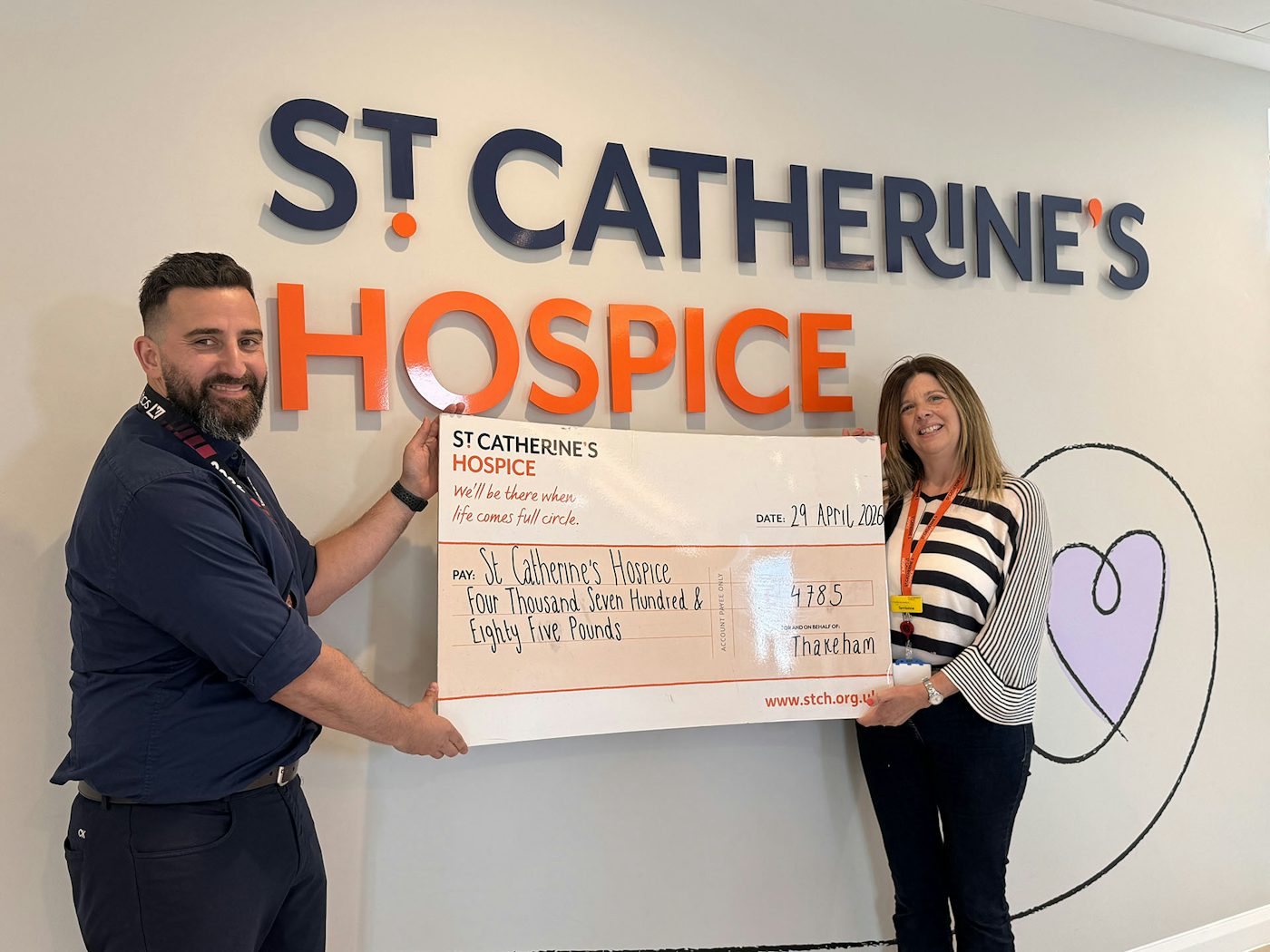Two people holding a large donation cheque for St Catherine’s Hospice in front of a wall sign reading ‘St Catherine’s Hospice’