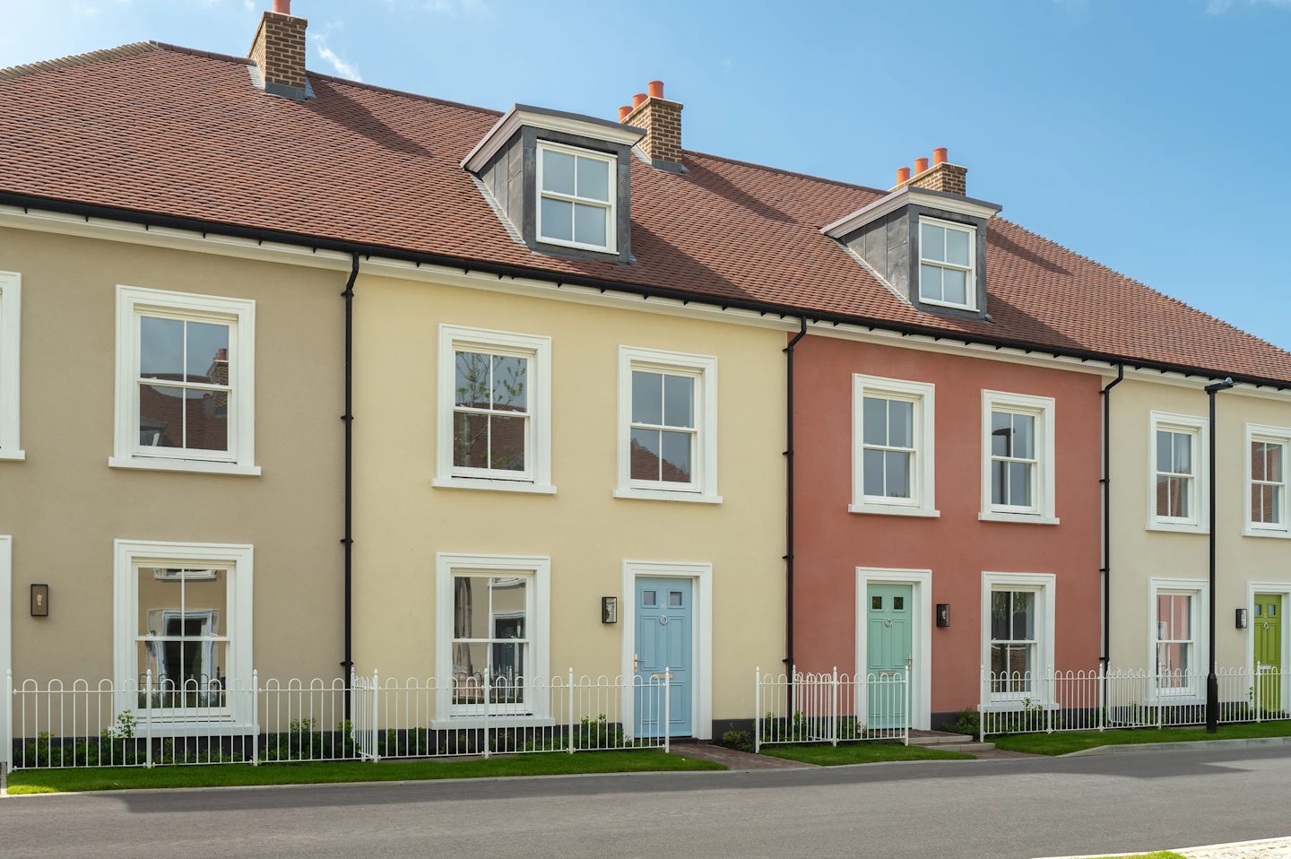 Row of terraced houses with cream and red walls, white windows, and pastel coloured front doors behind a low white fence