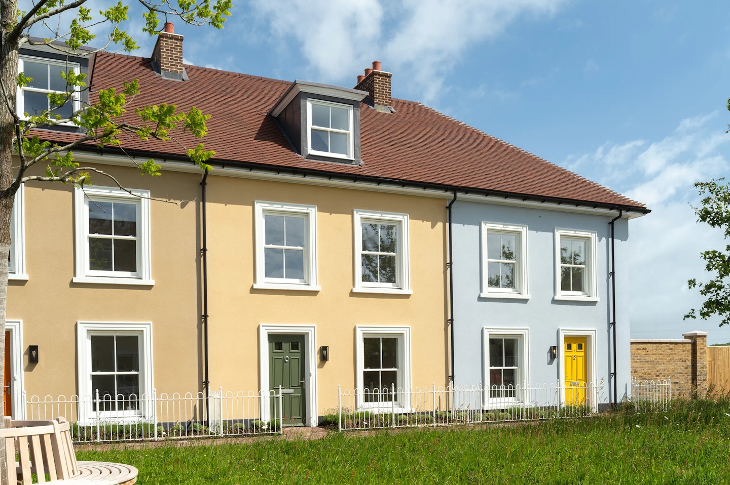Row of terraced houses with cream and light blue walls, coloured front doors, and grass and a small bench in front