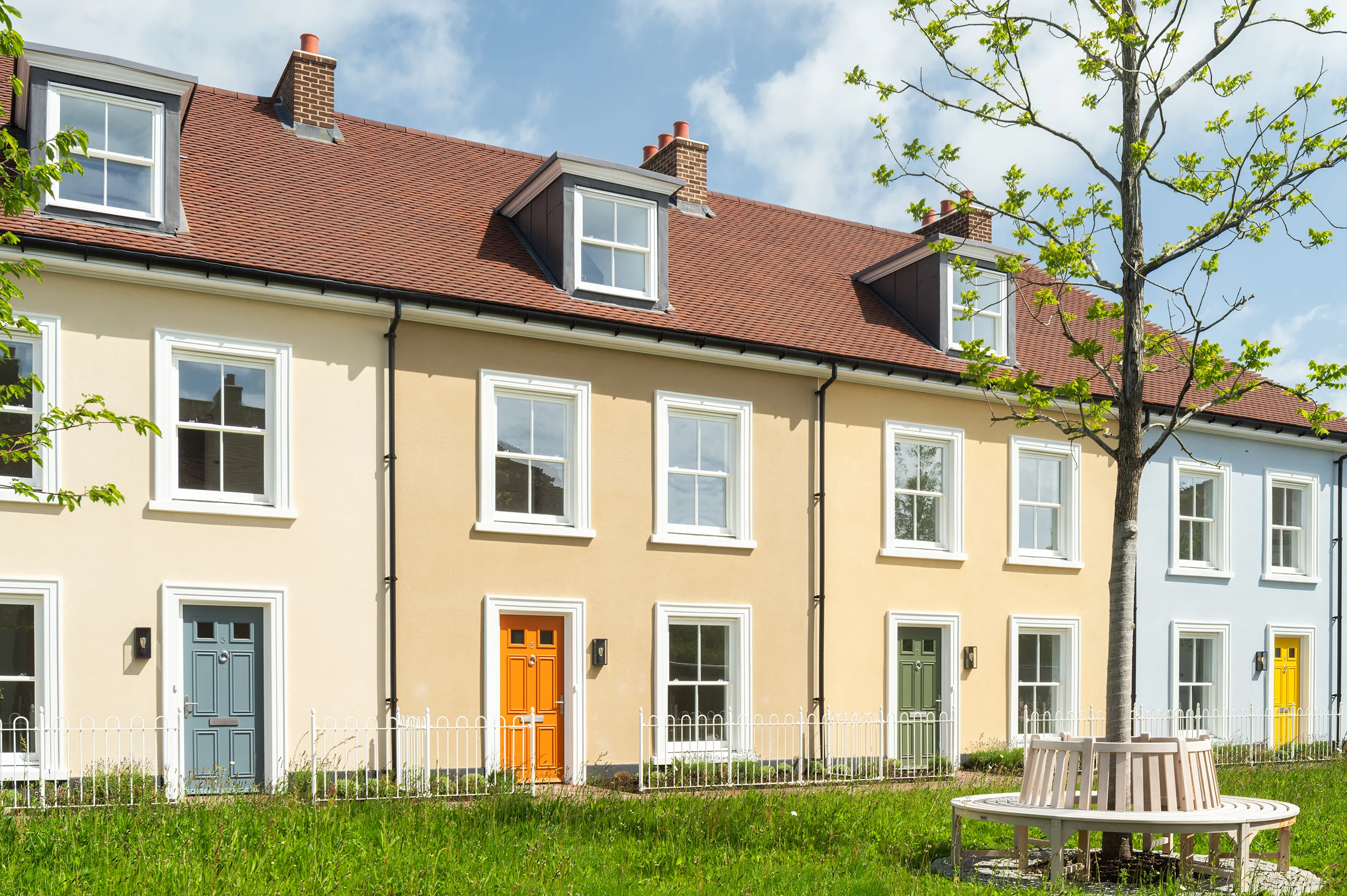 Terraced houses with cream and light blue walls, coloured front doors, and a tree with a circular bench in the foreground
