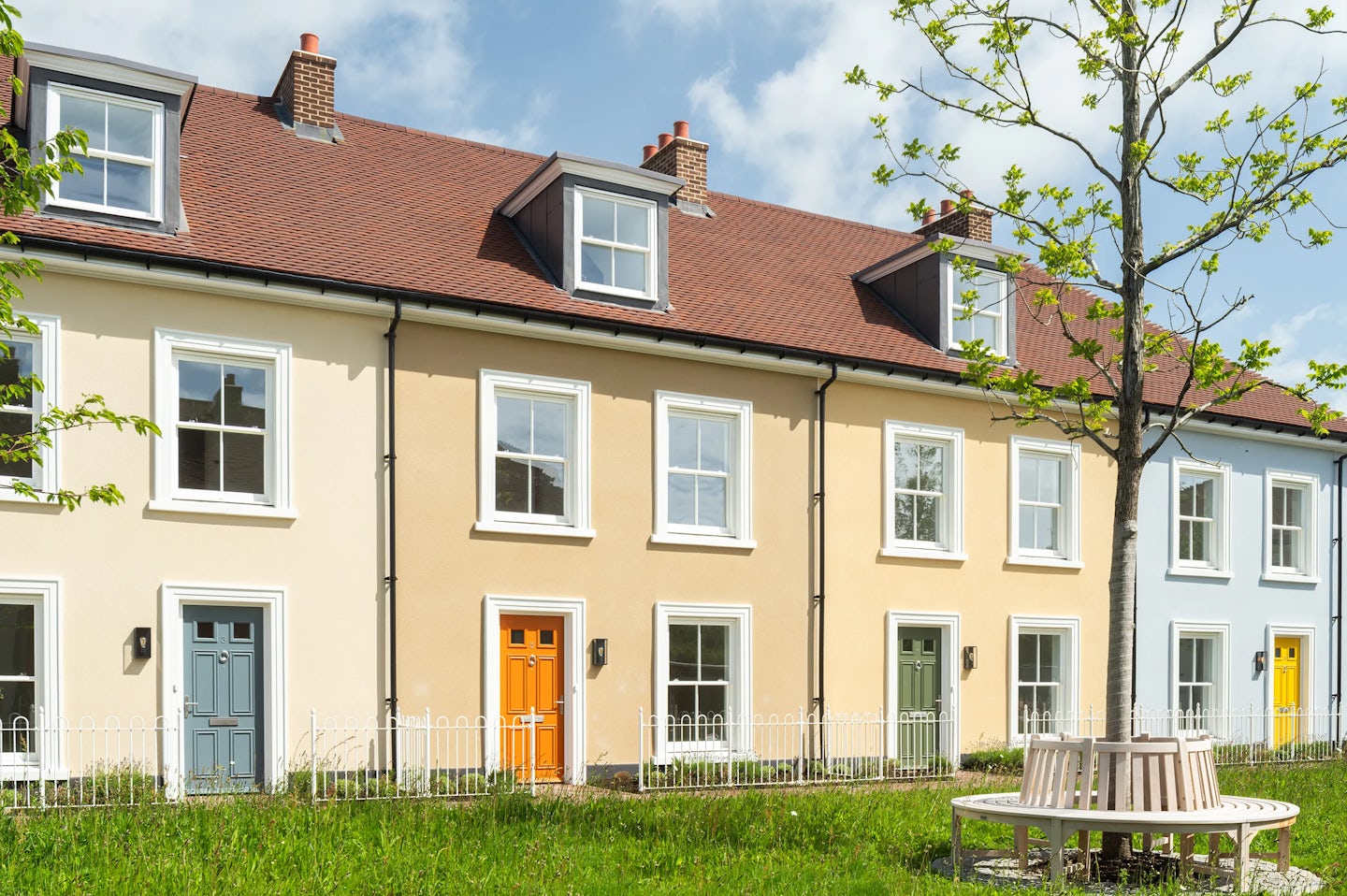 Terraced houses with cream and light blue walls, coloured front doors, and a tree with a circular bench in the foreground