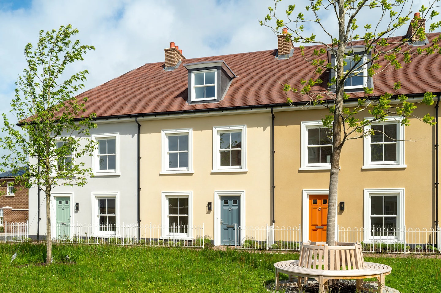 Row of two-storey terraced houses with cream and white walls, coloured front doors, and a small tree and bench in front