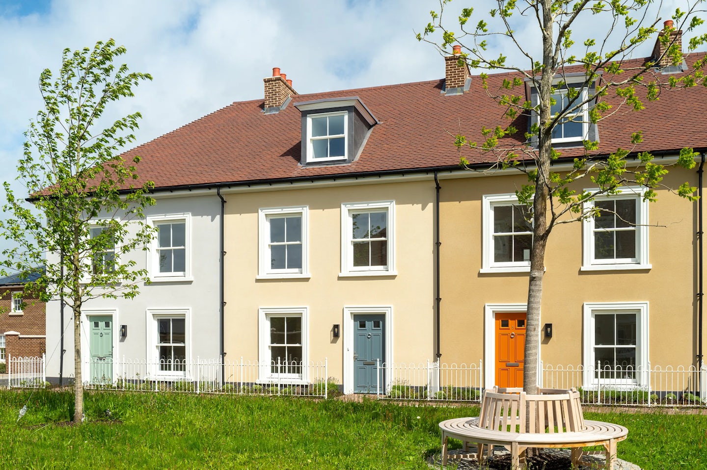Row of two-storey terraced houses with cream and white walls, coloured front doors, and a small tree and bench in front