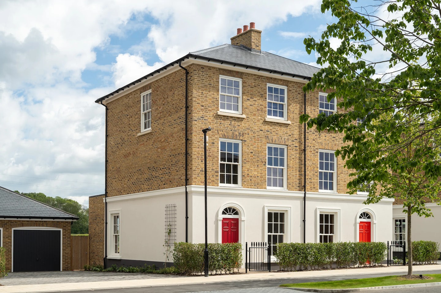 Three-storey brick townhouse with white ground floor and red front doors, with hedges and a gate along the pavement