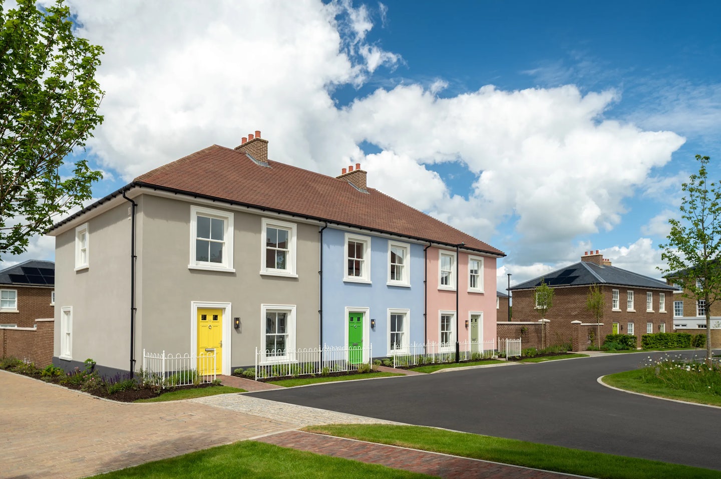 Row of pastel coloured houses with white windows and coloured front doors along a curved residential street with trees and grass