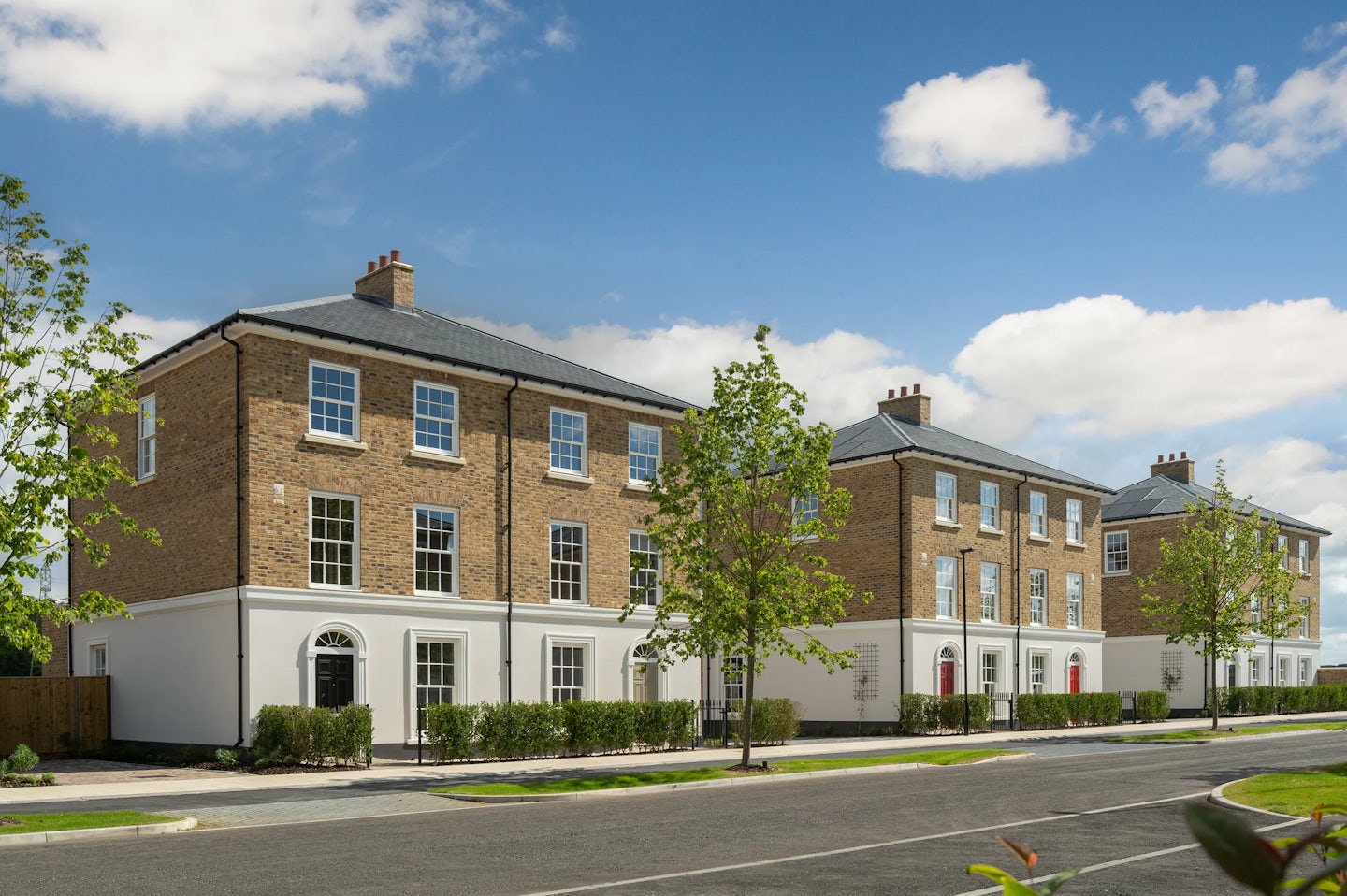 Large three-storey brick townhouses with white ground floor, trees and hedges along a quiet road