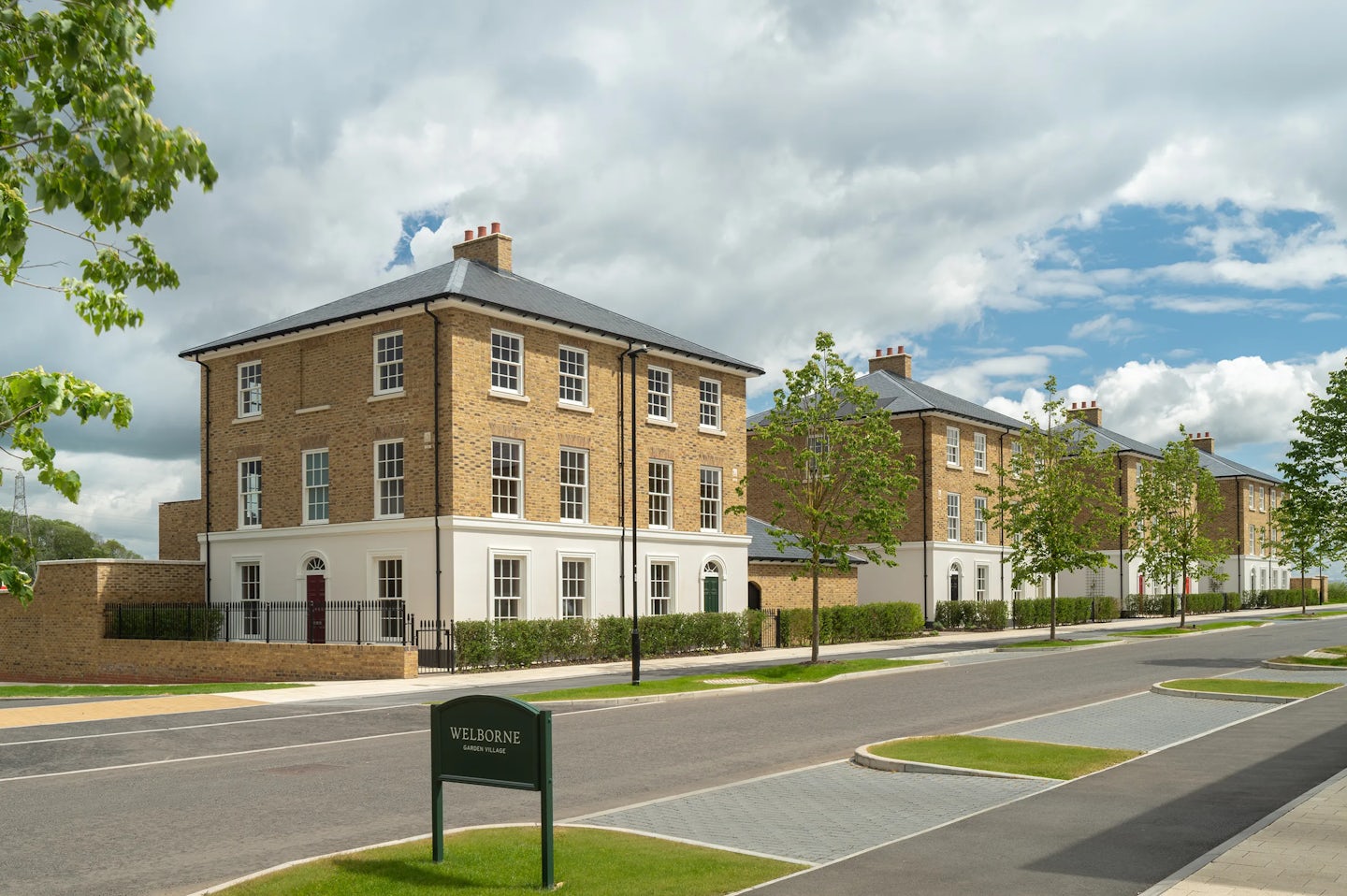 Large three-storey brick townhouses with white ground floor, trees and hedges along a quiet road